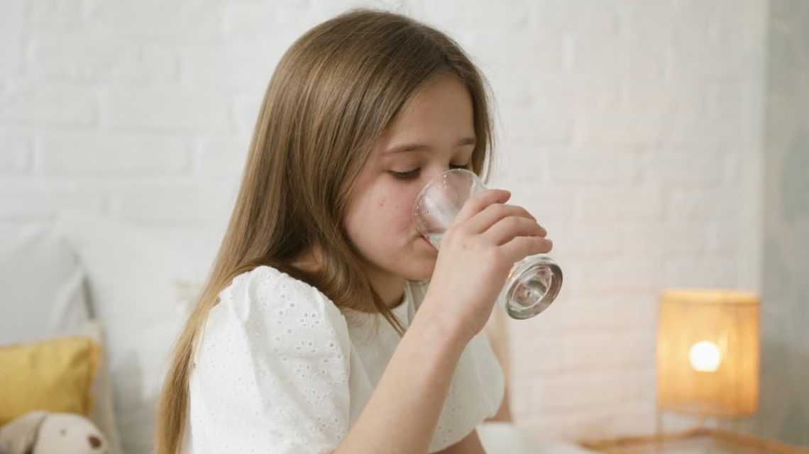 A young girl drinks water in a cozy bedroom, highlighting hydration and comfort.