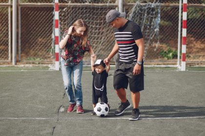 Full body of positive happy parents in casual clothes holding toddler son hands while walking together on football field on sunny day